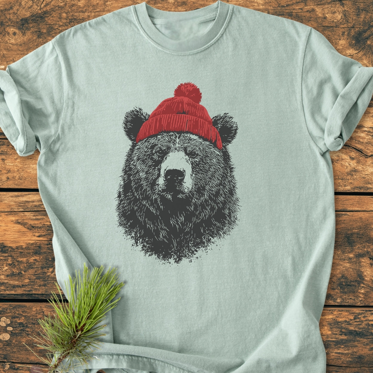 Bear With Beanie - Vintage Wash Tee features a detailed illustration of a bear wearing a knit beanie, printed on a light-colored, soft-washed, relaxed fit T-shirt, laid on a wooden surface.