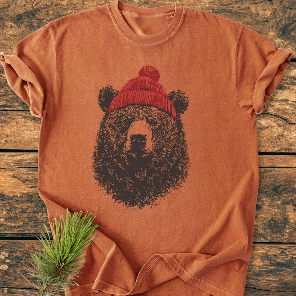 Bear With Beanie - Vintage Wash Tee featuring a stylized bear's head with a red knit beanie, laid on wood with a pine sprig, showcases a relaxed fit and durable stitching.