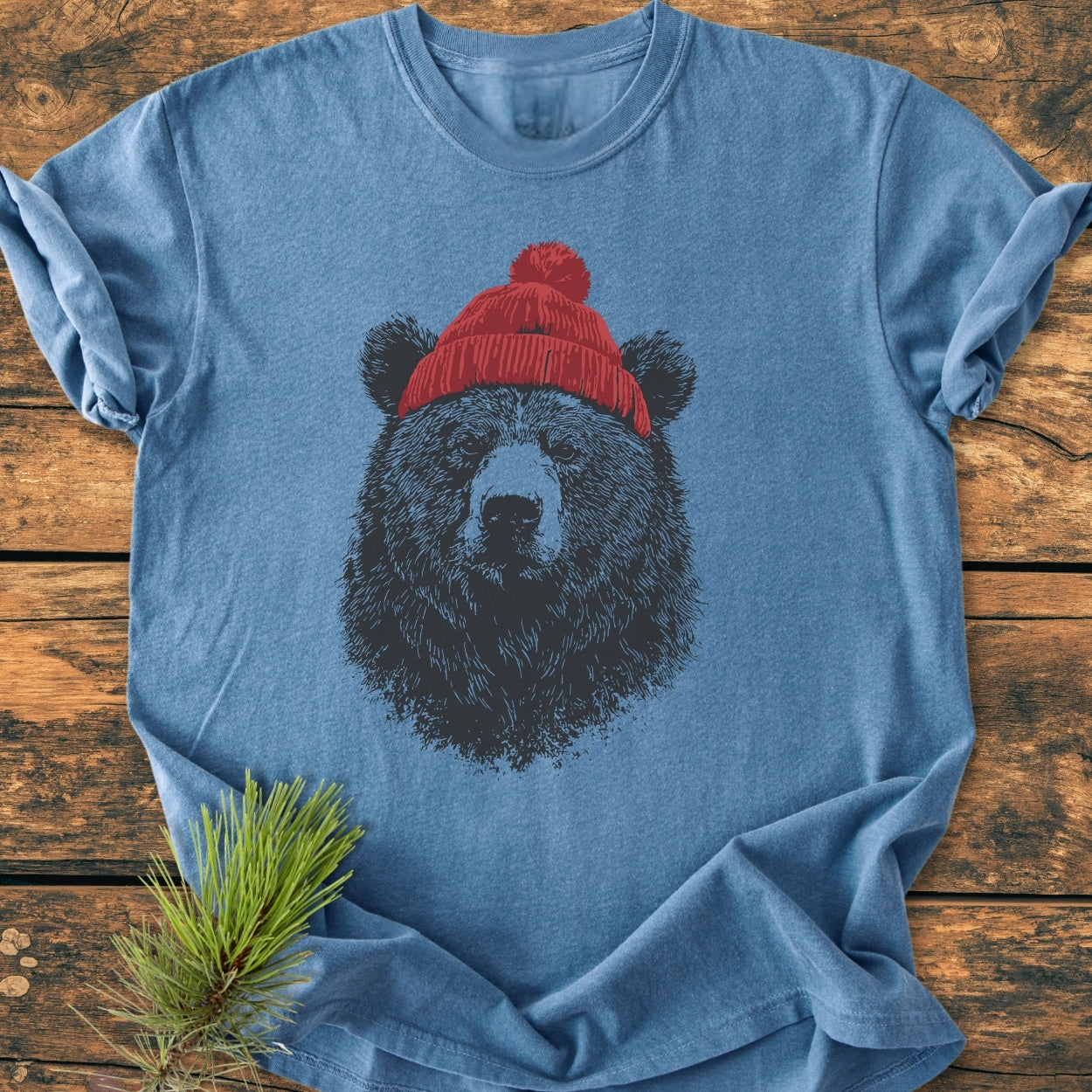 Bear With Beanie - Vintage Wash Tee featuring a black bear face in a red knit beanie, styled on a wooden surface with a pine branch, showcasing its relaxed fit and soft texture.