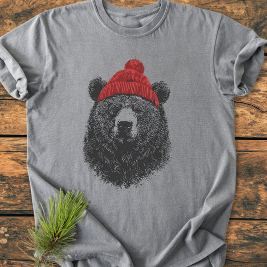 Bear With Beanie - Vintage Wash Tee features a black bear illustration with a red knit beanie on a heather-gray T-shirt, laid flat on a rustic wooden surface with a pine sprig accent.