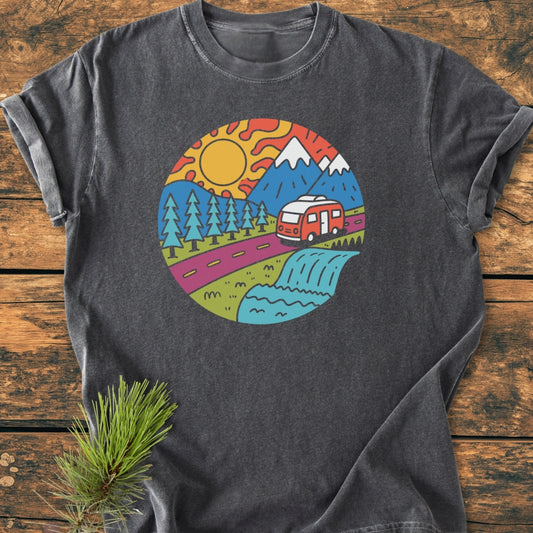 Scenic Route - Vintage Wash Tee featuring a colorful road-trip scene with mountains, pine trees, a red camper van, and a waterfall. Laid on a wooden surface with a pine sprig accent.