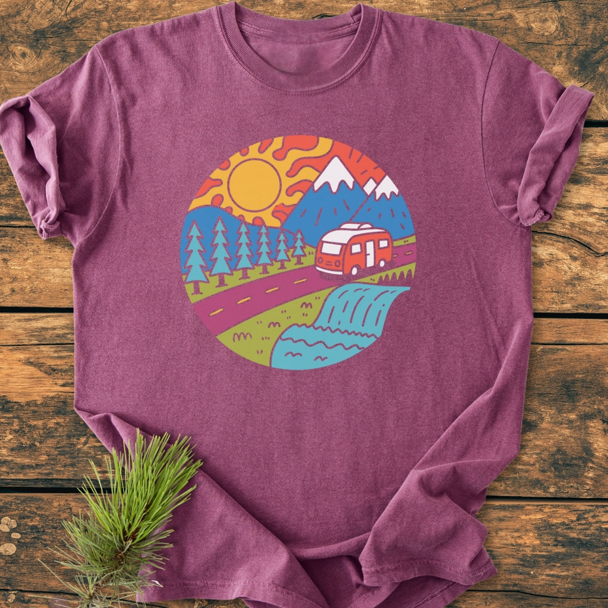 Scenic Route - Vintage Wash Tee: Maroon-purple T-shirt displaying a cartoon nature scene with mountains, sun, trees, road, camper van, and river. Laid flat, highlighting its soft, relaxed fit and durable stitching.