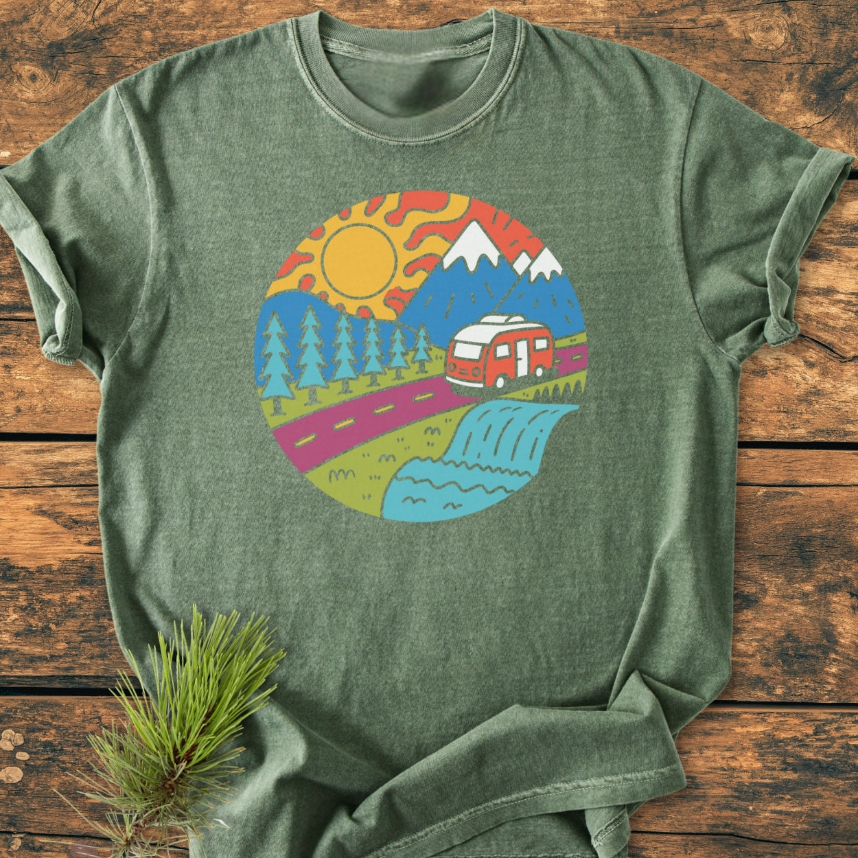 Scenic Route - Vintage Wash Tee in heather-green, featuring a colorful outdoor road-tripping graphic with mountains, trees, and a camper, displayed on rustic wood with a pine sprig beside it.