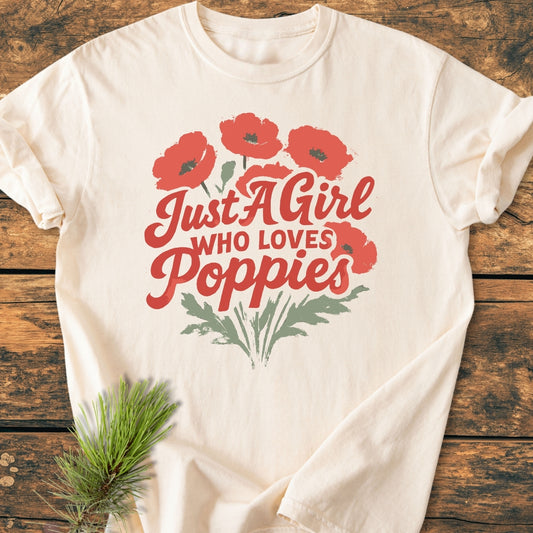 Just A Girl Who Loves Poppies - Vintage Wash Tee featuring red poppy graphics and text on an off-white T-shirt, laid flat on a wooden surface, accented by a sprig of greenery.