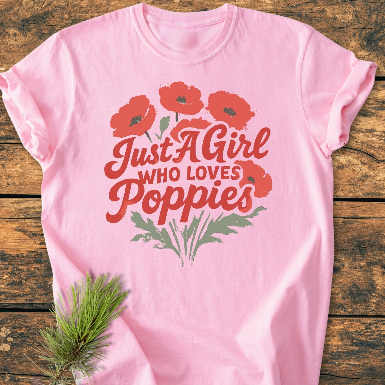 Just A Girl Who Loves Poppies - Vintage Wash Tee features red poppy graphics and text on a soft pink background, laid flat on wood with a pine sprig accenting the scene.