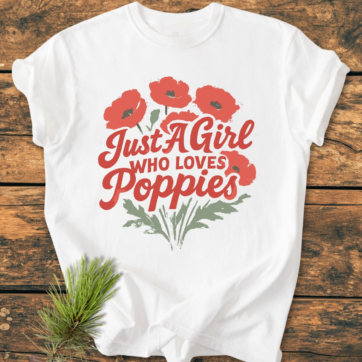 The Just A Girl Who Loves Poppies - Vintage Wash Tee features red poppies and green stems on a white background, laid on wood, complemented by a pine sprig.