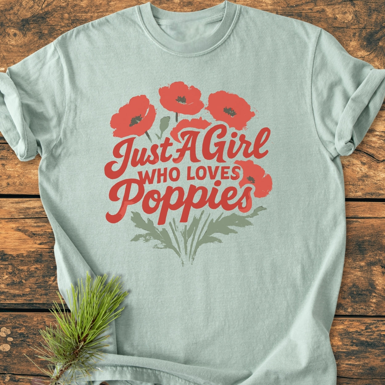 Just A Girl Who Loves Poppies - Vintage Wash Tee features red poppy bouquet design on pale mint-green fabric. Laid flat on wood, styled with a pine sprig for a cozy, artisanal feel.