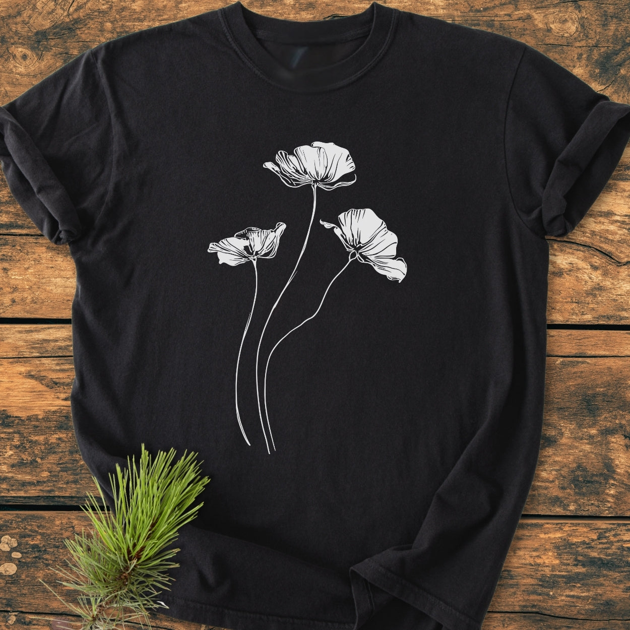Delicate Poppies - Vintage Wash Tee features white line art poppy flowers on a black T-shirt, laid on a wooden surface. Made of soft-washed, garment-dyed cotton for a comfortable, relaxed fit.