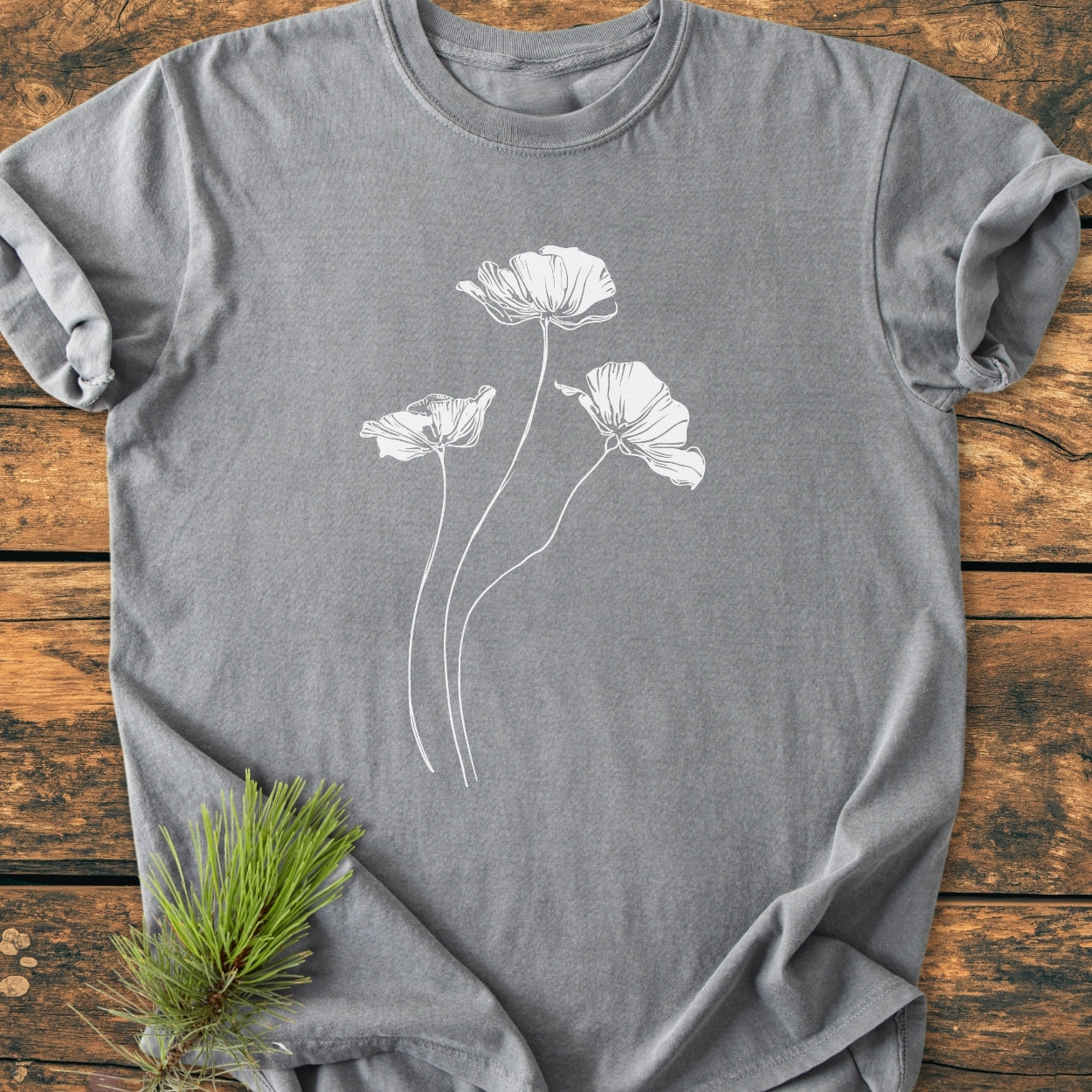 Delicate Poppies - Vintage Wash Tee features a heather-gray T-shirt with a white line-drawing of poppy-like flowers, laid flat on wood, accented by a pine sprig, showcasing its relaxed, cozy design.