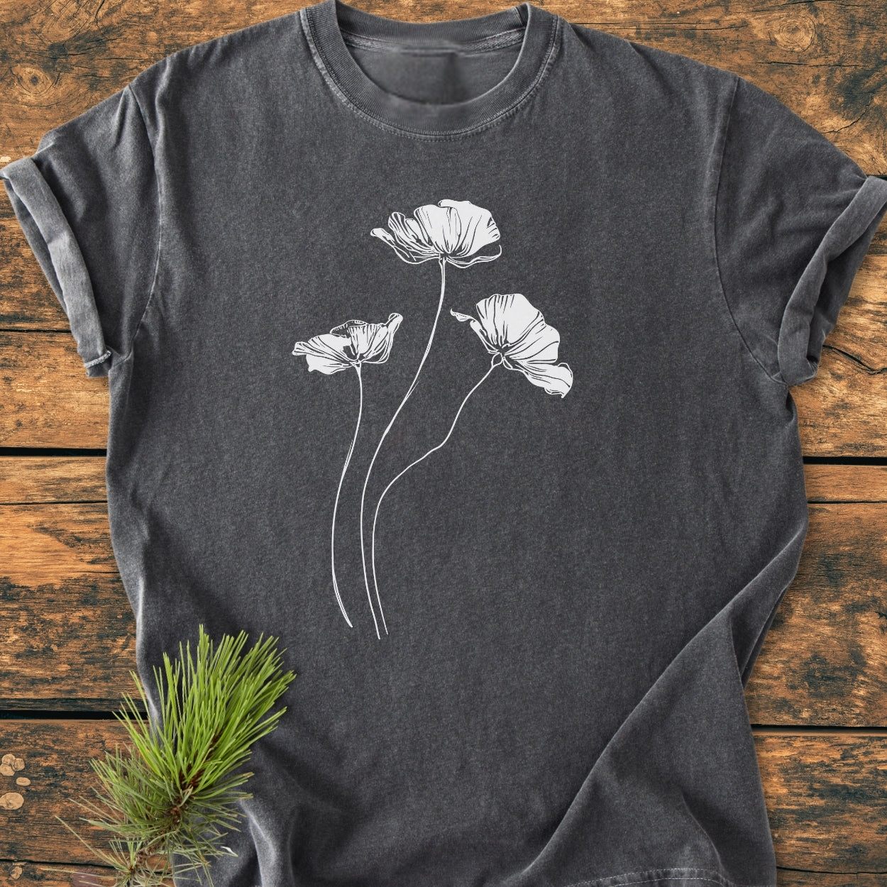 Delicate Poppies - Vintage Wash Tee featuring a white line-drawing of poppy-style flowers on a dark heather-gray T-shirt, styled with a rustic pine sprig on a wooden surface.