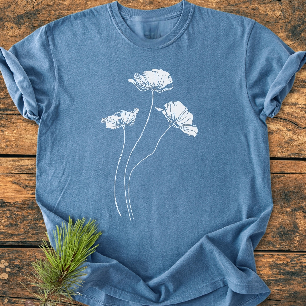 Delicate Poppies - Vintage Wash Tee in heather-blue with white poppy line-drawing, laid flat on wooden surface, accompanied by a pine sprig, showcasing its relaxed fit and garment-dyed texture.