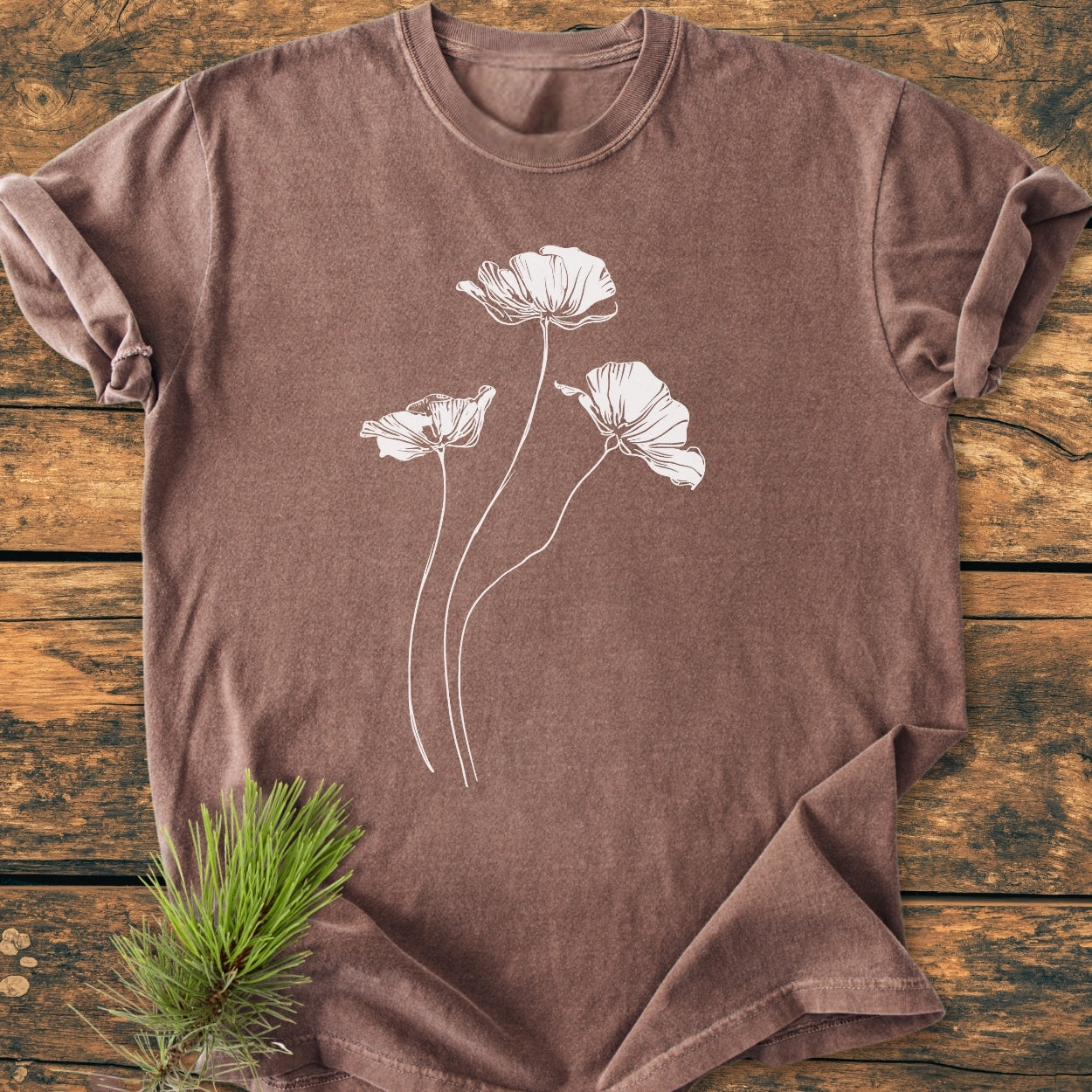 Delicate Poppies - Vintage Wash Tee featuring white poppy-style flowers on a brown short-sleeved shirt, displayed on a wooden surface with a pine sprig beside it, highlighting its rustic charm.
