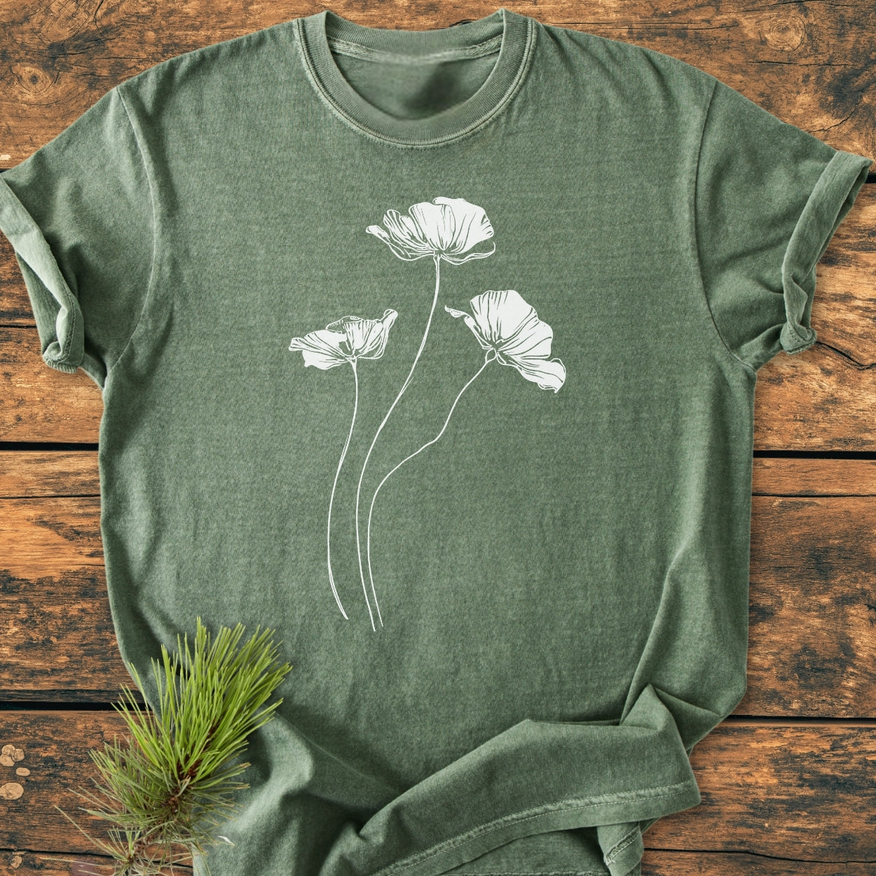 Delicate Poppies - Vintage Wash Tee in heather-green with white poppy line art, laid flat on wood. A pine sprig accents the soft, relaxed-fit, garment-dyed Comfort Colors shirt.