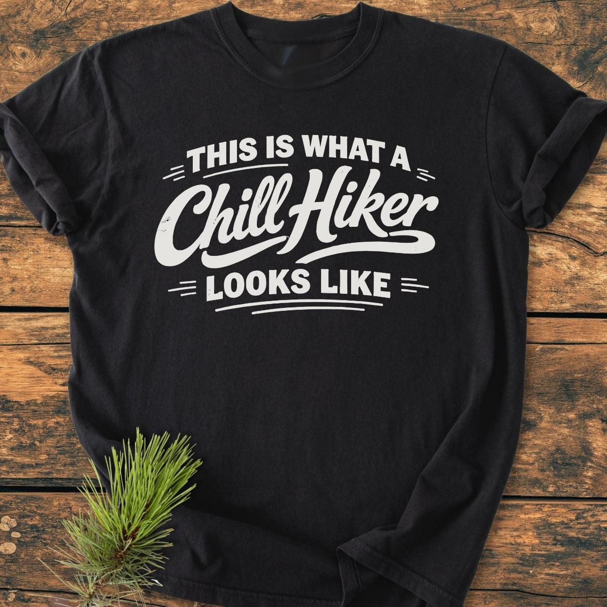 Chill Hiker - Vintage Wash Tee with This Is What A Chill Hiker Looks Like in white text, laid flat with a pine sprig accent on the wooden surface.