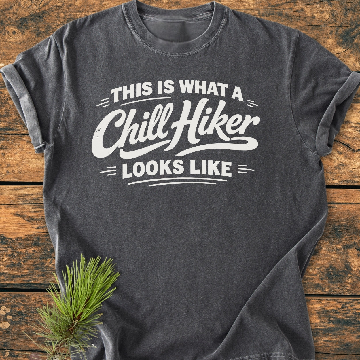 Chill Hiker - Vintage Wash Tee in charcoal-gray, featuring bold white lettering on chest. Laid flat with rolled sleeves on wooden surface, pine sprig beside, highlighting its outdoor theme.