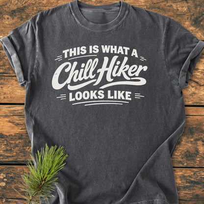 Chill Hiker - Vintage Wash Tee in charcoal-gray, featuring bold white lettering on chest. Laid flat with rolled sleeves on wooden surface, pine sprig beside, highlighting its outdoor theme.