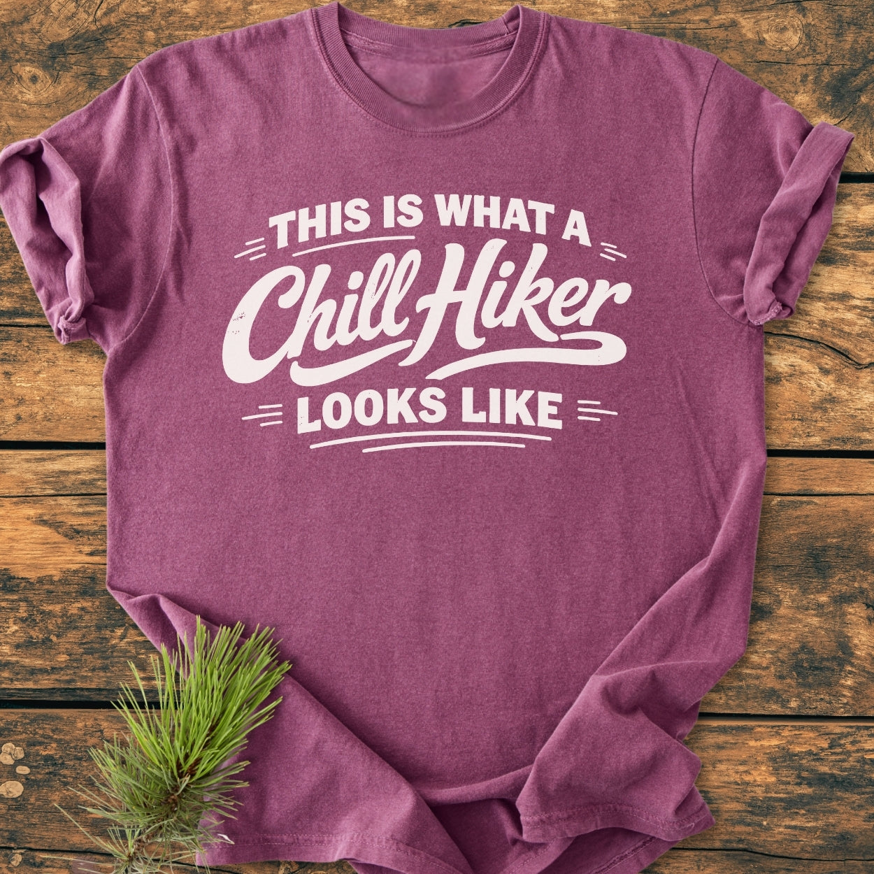 Chill Hiker - Vintage Wash Tee in heathered purple/mauve, laid flat on wood-grain surface. Features white slogan text. Accented by a sprig of pine, showcasing relaxed fit and soft-washed fabric.
