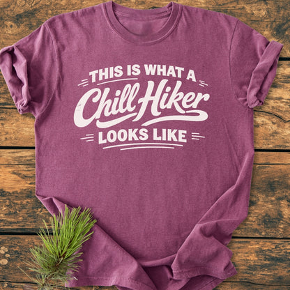 Chill Hiker - Vintage Wash Tee in heathered purple/mauve, laid flat on wood-grain surface. Features white slogan text. Accented by a sprig of pine, showcasing relaxed fit and soft-washed fabric.
