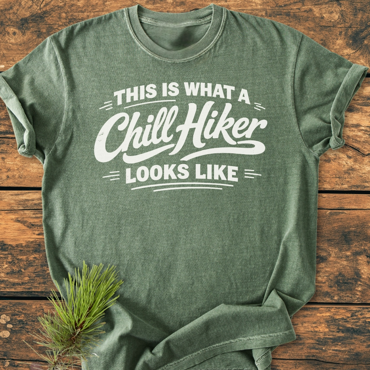 Chill Hiker - Vintage Wash Tee in heather-green, featuring a This is what a Chill Hiker looks like slogan, displayed folded on wood with a pine sprig beside it.