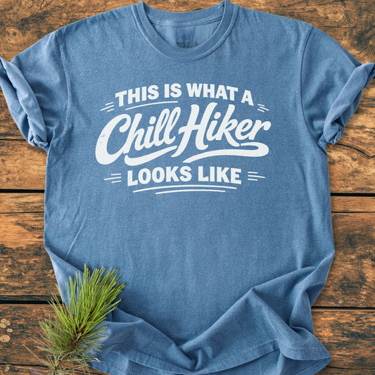 Chill Hiker - Vintage Wash Tee in heather-blue, featuring bold white text, laid flat on rustic wood. Accompanied by a pine sprig, emphasizing its outdoorsy appeal and relaxed fit.