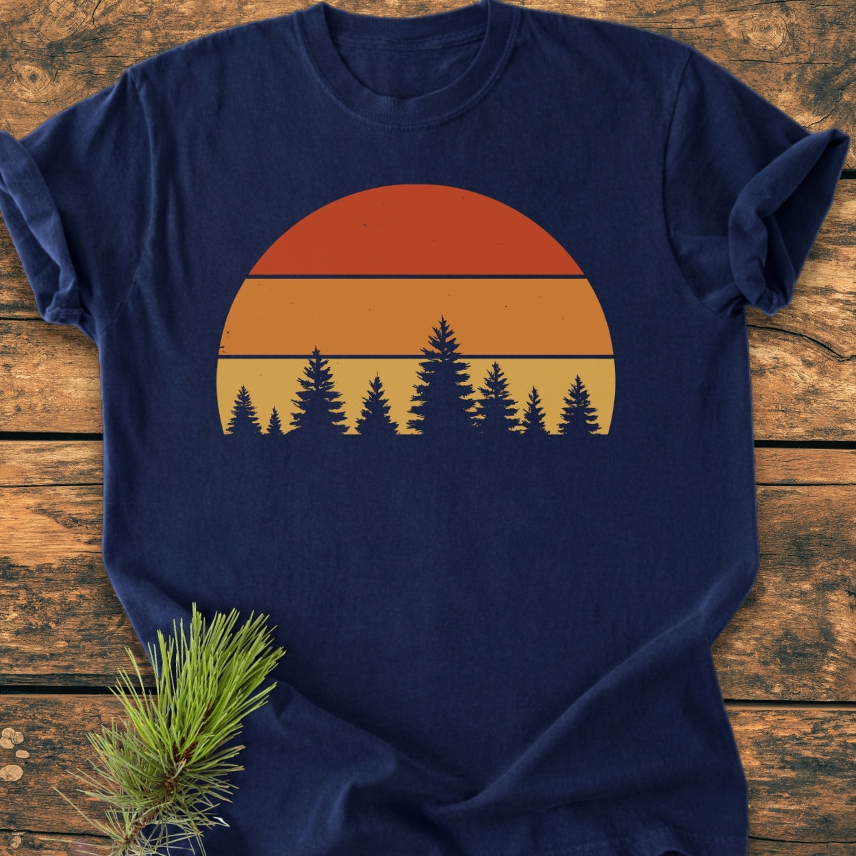 Forest Sunset - Vintage Wash Tee: Navy blue t-shirt featuring a half-sunset design over pine trees, displayed on rustic wood with pine needles. Garment-dyed, ring-spun cotton for comfort and durability.