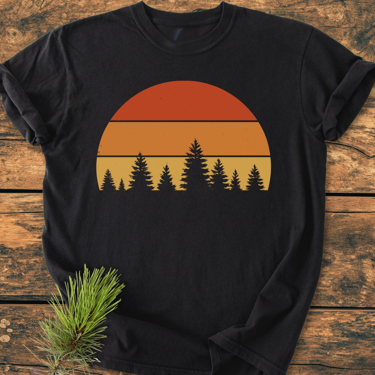 Forest Sunset - Vintage Wash Tee features a circular sunset graphic with pine-tree silhouettes on a black T-shirt, laid flat on a wooden surface, highlighting its relaxed, cozy, and durable design.