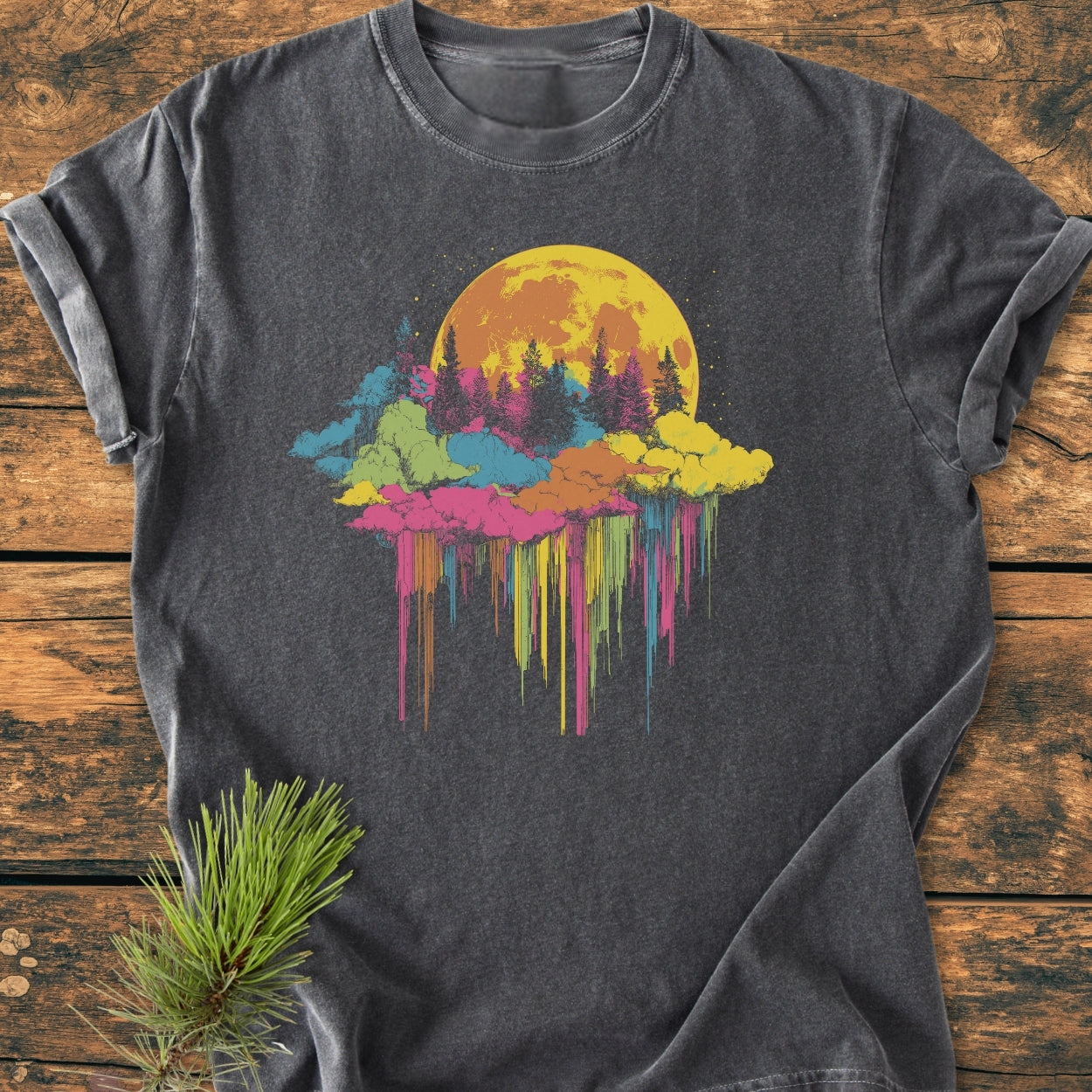 Moonlight Meltdown - Vintage Wash Tee featuring a vibrant moon and pine tree graphic, laid flat on wood with a pine sprig. The charcoal-gray, soft-washed, garment-dyed fabric offers a relaxed, comfortable fit.