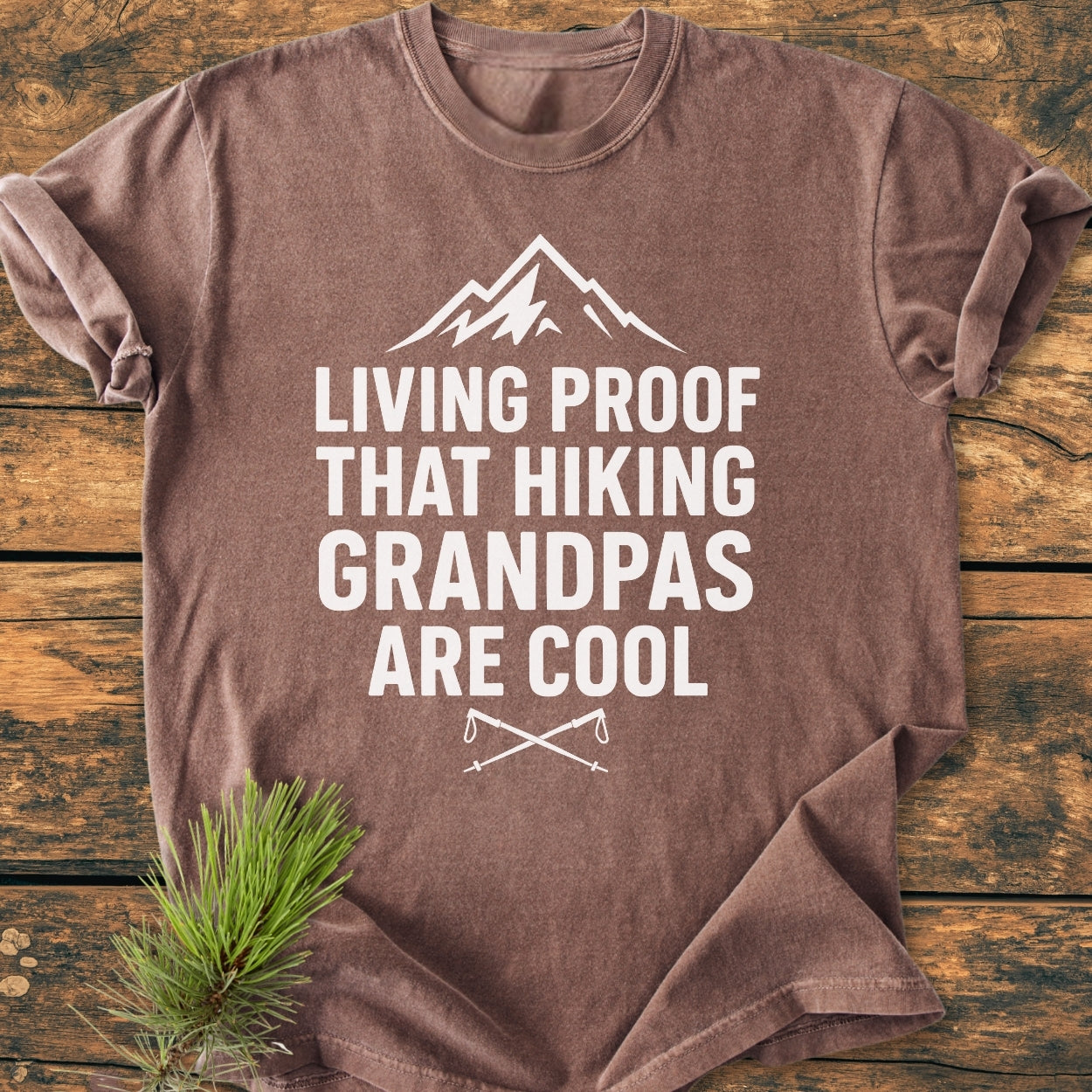 Living Proof Grandpa - Vintage Wash Tee, featuring a mountain outline and hiking slogan, is folded on a wooden surface with crossed hiking poles design and a pine sprig accentuating the outdoor theme.