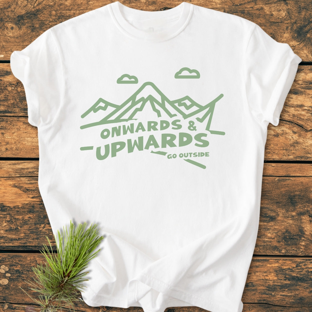 Go Outside - Vintage Wash Tee features a white crew-neck with sage-green line-art of mountains and slogan. Laid flat on wood, it includes a pine sprig prop, emphasizing its relaxed fit and durability.