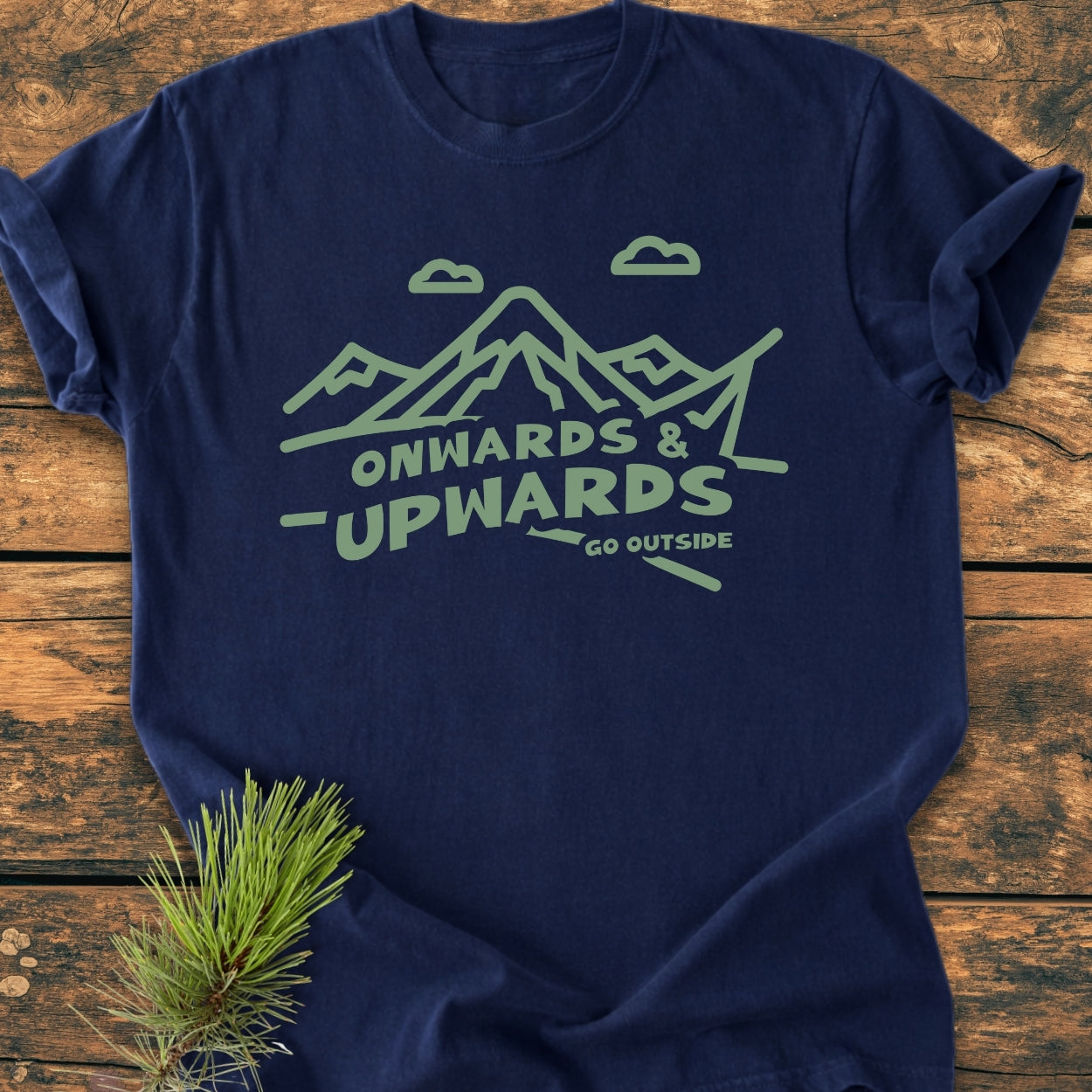 Go Outside - Vintage Wash Tee features a minty green mountain graphic and text on a navy-blue, garment-dyed fabric, laid on wood, paired with a pine branch to emphasize its outdoor theme.