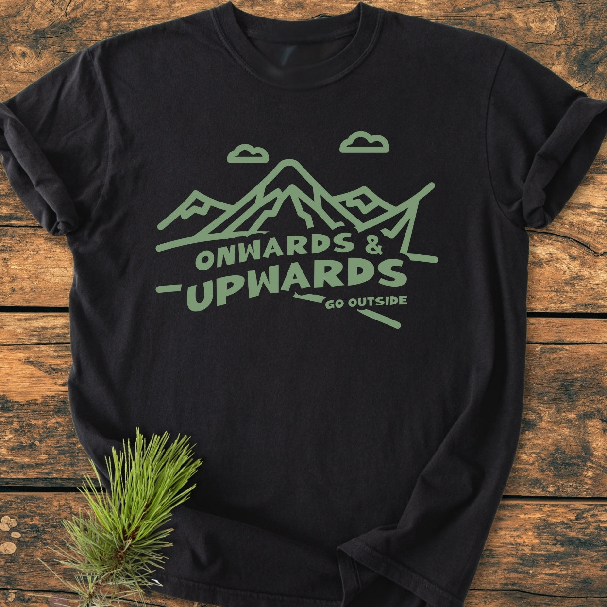 Go Outside - Vintage Wash Tee features a black crew-neck with a moss-green mountain design and slogan. Laid on wood, it embodies adventure, enhanced by a pine branch accent.