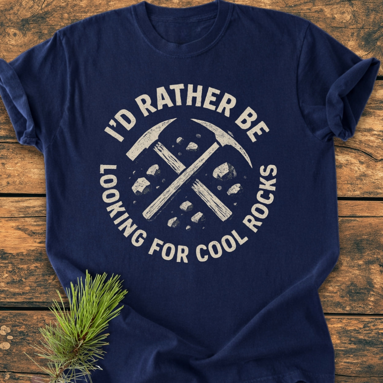 I'd Rather... Rockhound - Vintage Wash Tee laid on wood, features crossed rock-hammers with stones design, styled with a pine branch, showcasing its relaxed fit and soft texture.