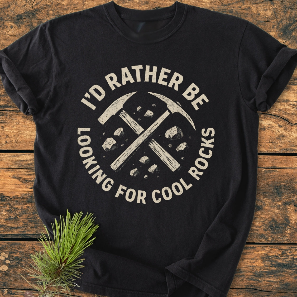 I’d Rather... Rockhound - Vintage Wash Tee features a circular rock-hammer design on a black Comfort Colors T-shirt, styled with a pine sprig, emphasizing its relaxed fit and durable, soft-washed fabric.