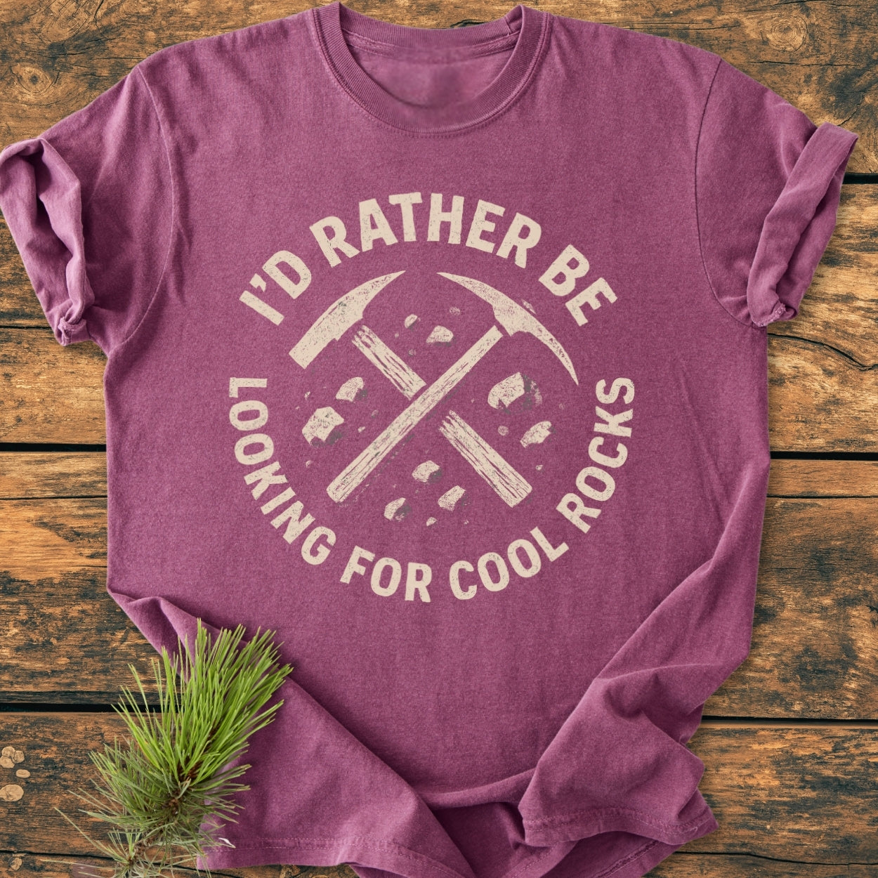 The I'd Rather... Rockhound - Vintage Wash Tee showcases a distressed graphic of crossed rock-hammers and stones on a cozy, heather-purple T-shirt, laid flat on a wooden surface.