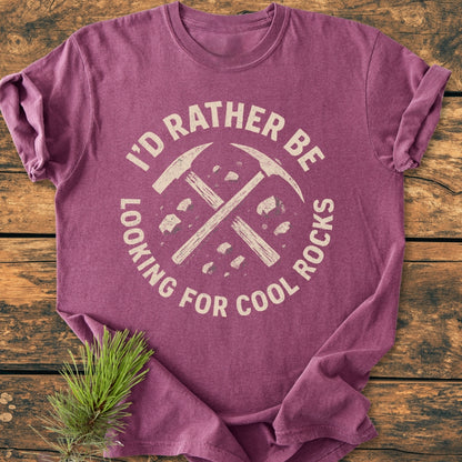 The I'd Rather... Rockhound - Vintage Wash Tee showcases a distressed graphic of crossed rock-hammers and stones on a cozy, heather-purple T-shirt, laid flat on a wooden surface.