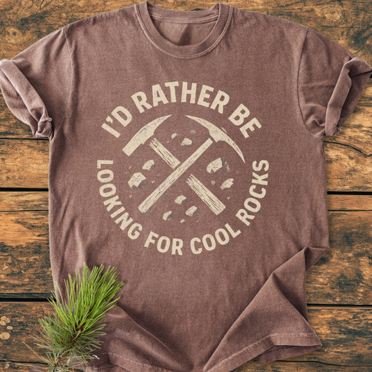 I'd Rather... Rockhound - Vintage Wash Tee features a brown cotton fabric with a distressed slogan and geologist tools design, laid on rustic wood, highlighted by a pine sprig for an outdoorsy feel.