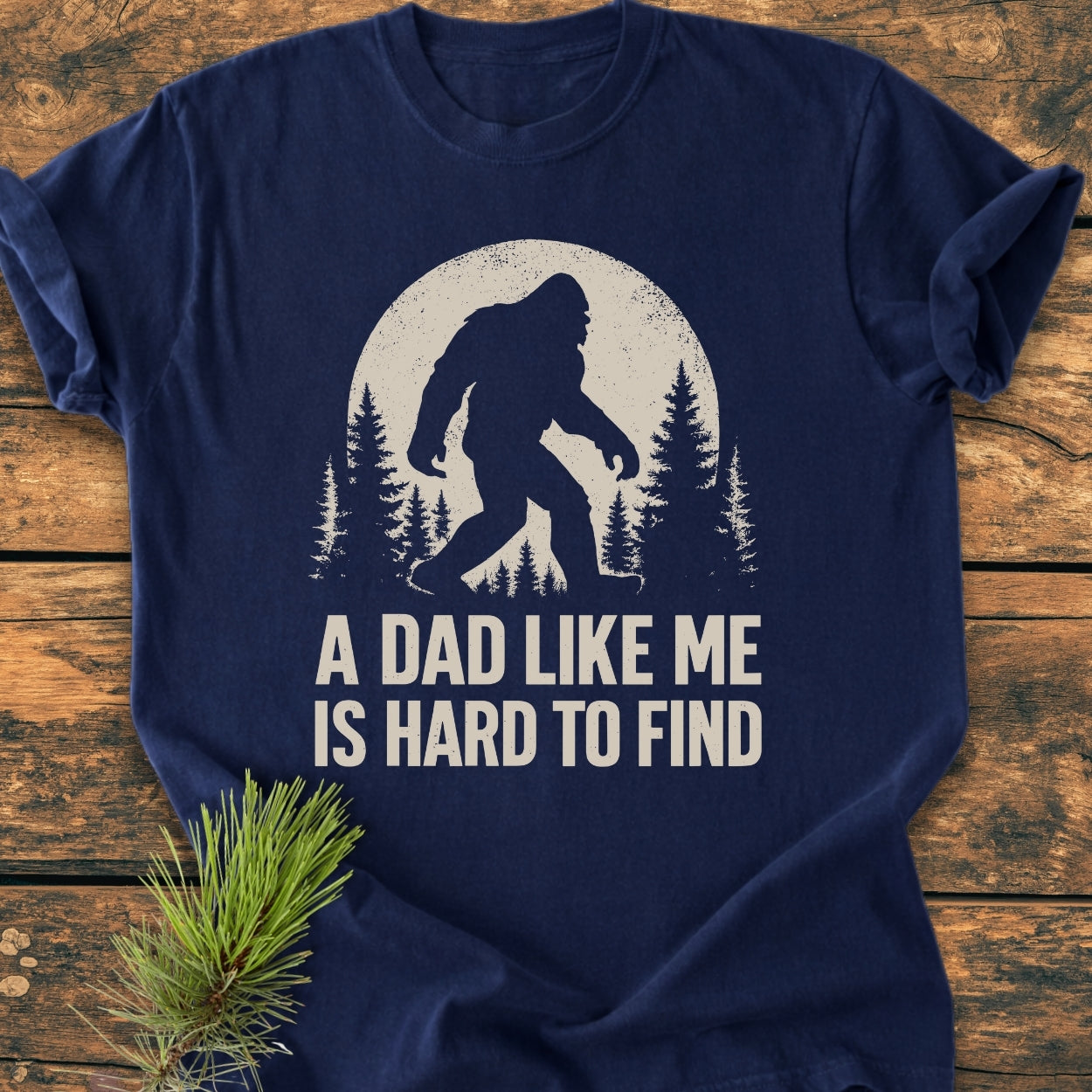 Vintage Bigfoot Dad - Vintage Wash Tee features a Sasquatch graphic with pine trees and moon. It includes the slogan “A DAD LIKE ME IS HARD TO FIND” on navy-blue cotton fabric.