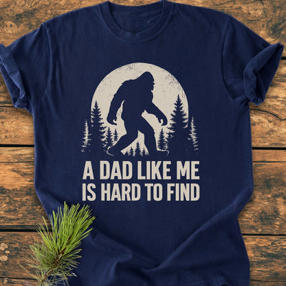 Vintage Bigfoot Dad - Vintage Wash Tee features a Sasquatch graphic with pine trees and moon. It includes the slogan “A DAD LIKE ME IS HARD TO FIND” on navy-blue cotton fabric.