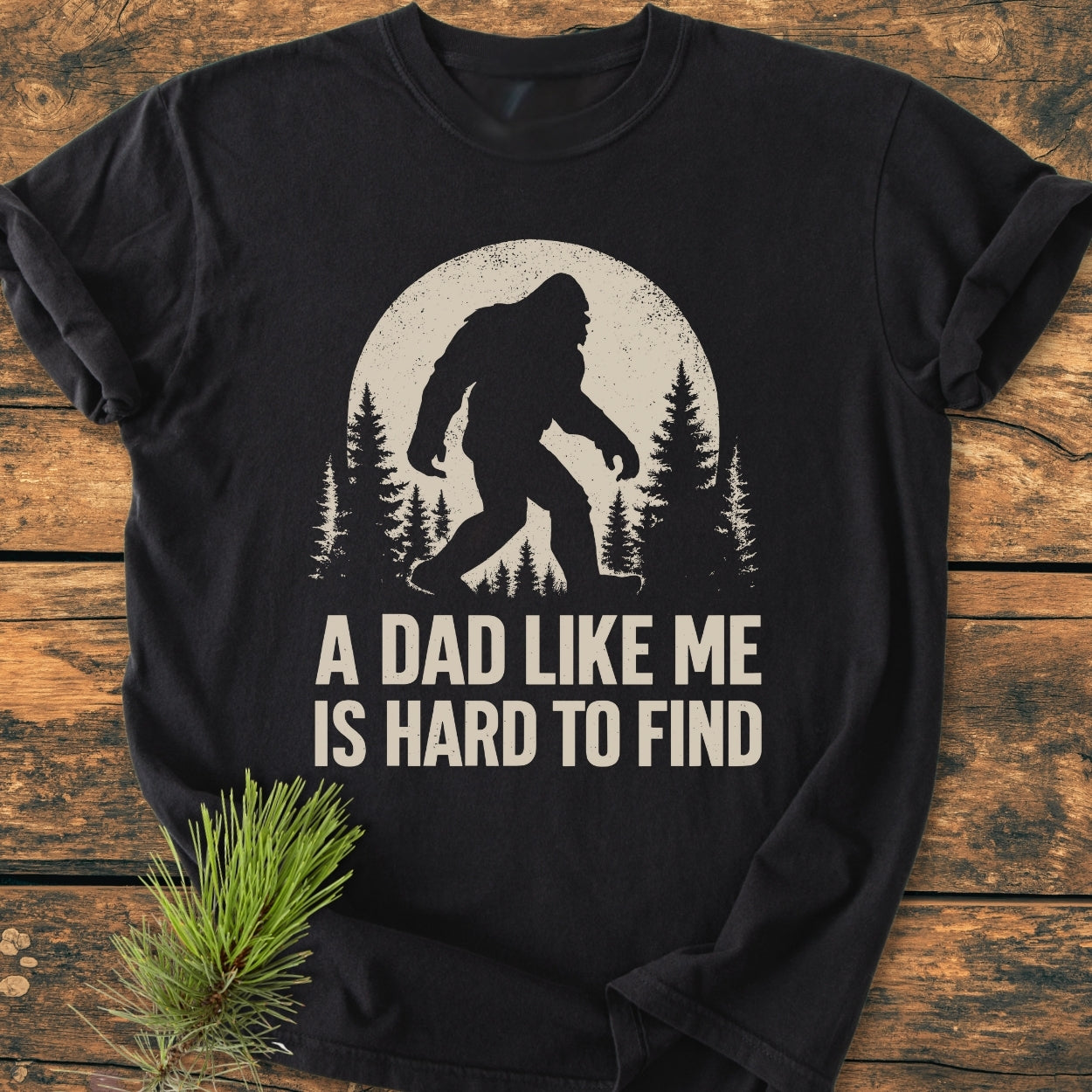 Vintage Bigfoot Dad - Vintage Wash Tee: Black T-shirt with Bigfoot silhouette and “A DAD LIKE ME IS HARD TO FIND” text, laid on wood with pine sprig decoration. Comfortable, relaxed fit, 100% cotton.