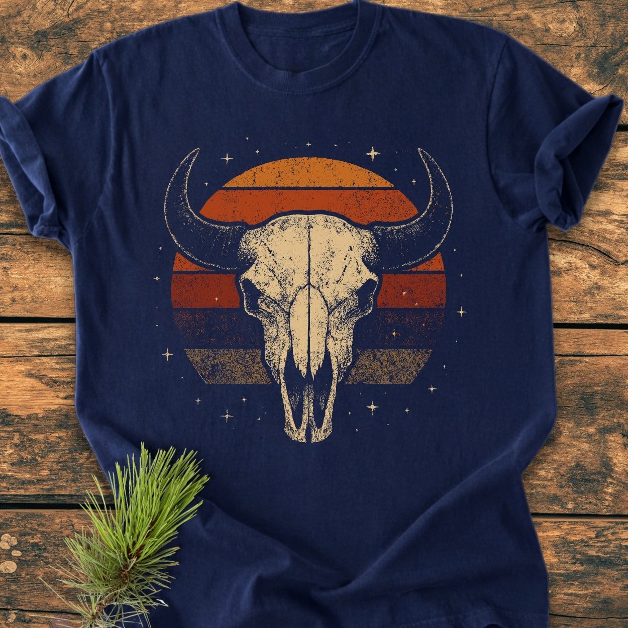 Retro Buffalo Skull - Vintage Wash Tee features a stylized buffalo skull with a retro sunset backdrop, printed on a navy-blue shirt laid on a wooden tabletop, with a pine sprig beside it.