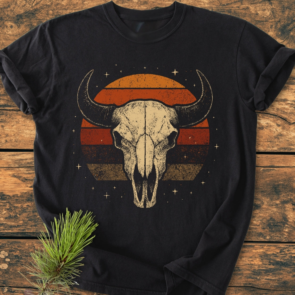 Retro Buffalo Skull - Vintage Wash Tee featuring a steer skull and sunset motif on black, laid on wood with pine twig accent, highlighting its soft, durable 100% ring-spun cotton construction.