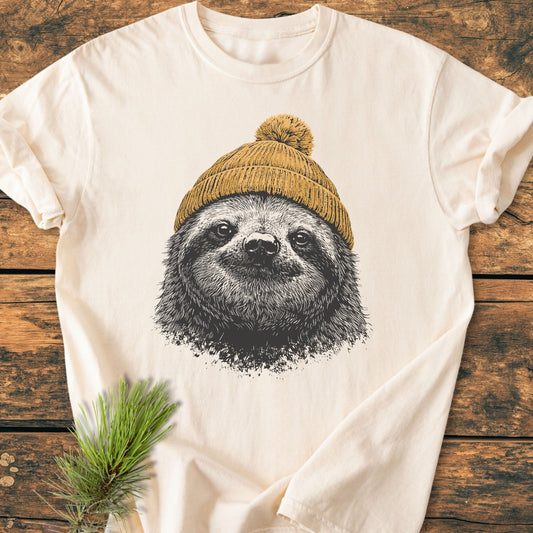 Sloth With Beanie - Vintage Wash Tee features a sloth face in a mustard beanie, printed on a cream T-shirt, laid on wood with a pine sprig for a cozy touch.