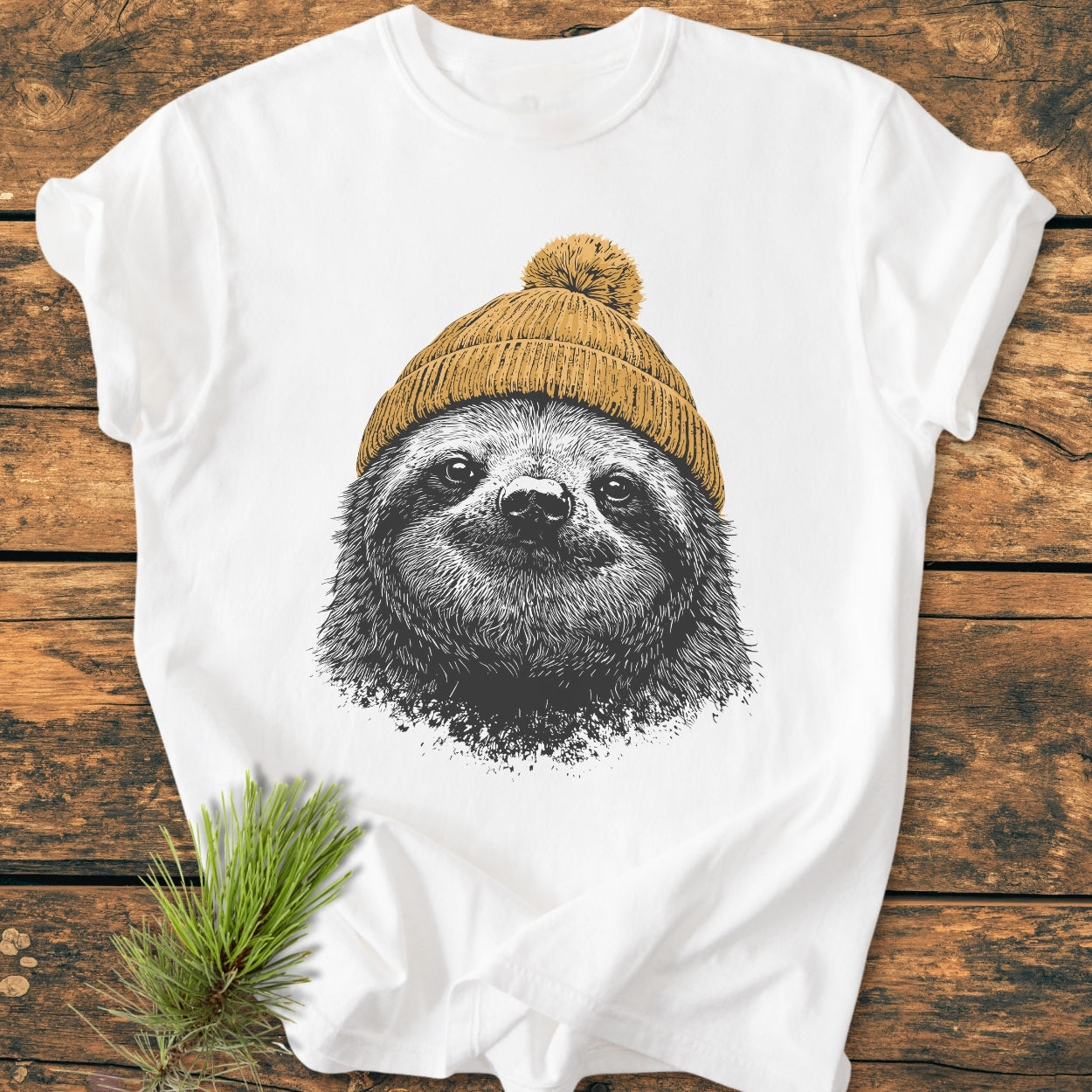 Sloth With Beanie - Vintage Wash Tee features a sloth sketch with a mustard-yellow beanie on a soft, garment-dyed white T-shirt, displayed on a rustic wooden surface with a pine branch nearby.