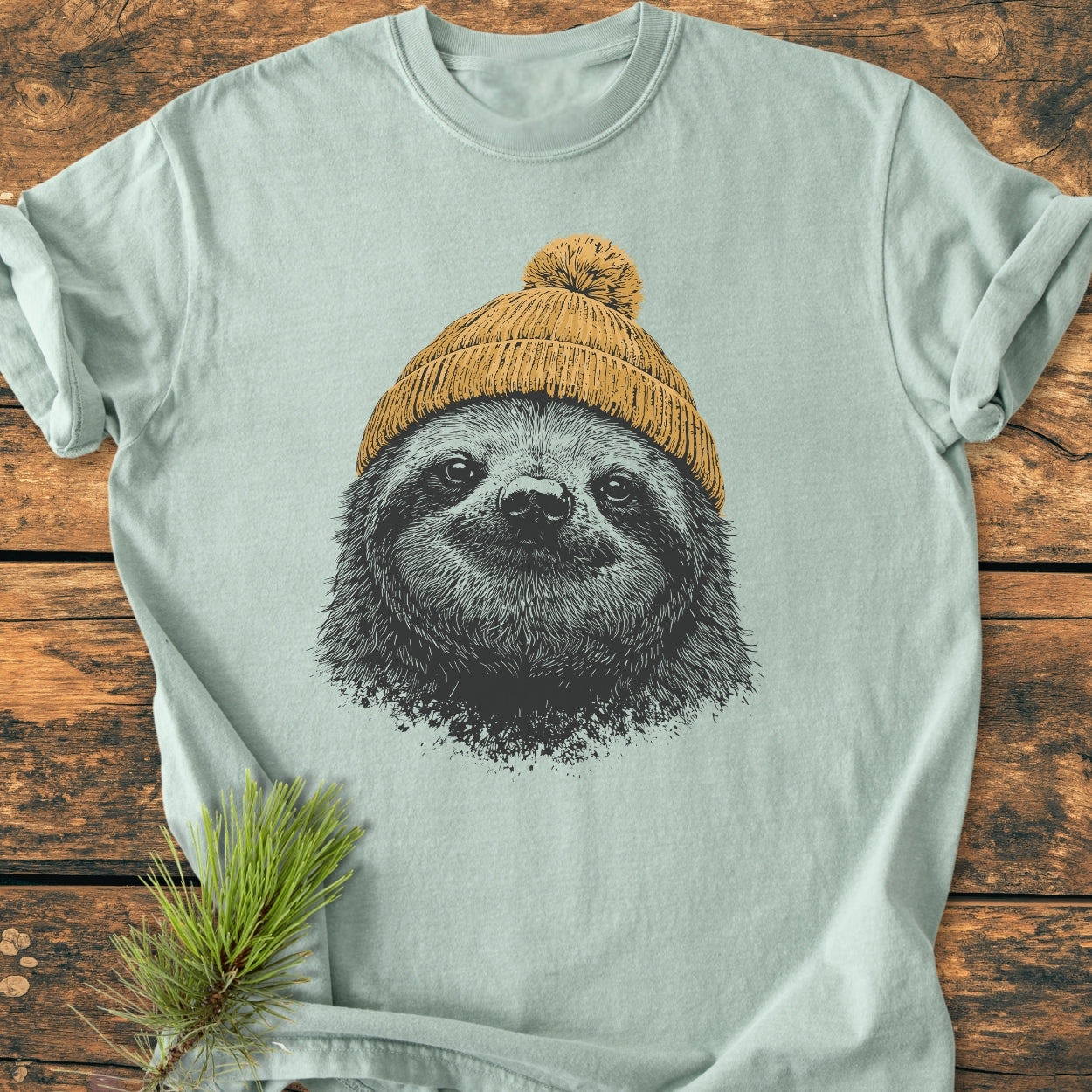 Sloth With Beanie - Vintage Wash Tee features a pastel green design with a sloth in a mustard-yellow beanie, laid on rustic wood, highlighting its relaxed fit and soft-washed texture.