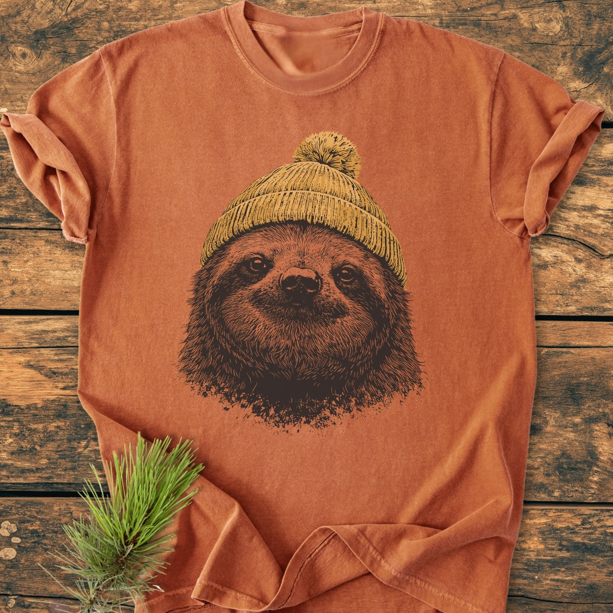 Sloth With Beanie - Vintage Wash Tee features a sloth face in a knit beanie design on a rust-orange short-sleeve shirt, laid on a wooden surface with a pine sprig accent.
