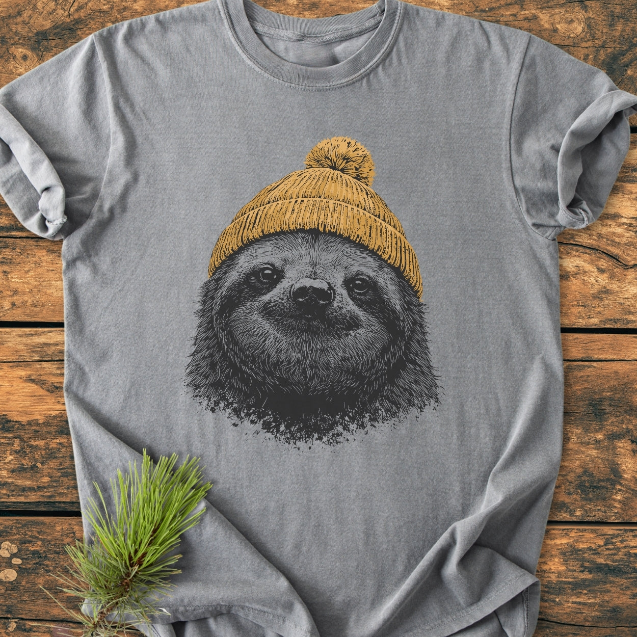 Sloth With Beanie - Vintage Wash Tee features a heather-grey T-shirt with a black-ink sloth face illustration wearing a mustard-yellow beanie, laid on a rustic wooden surface with a pine sprig accent.