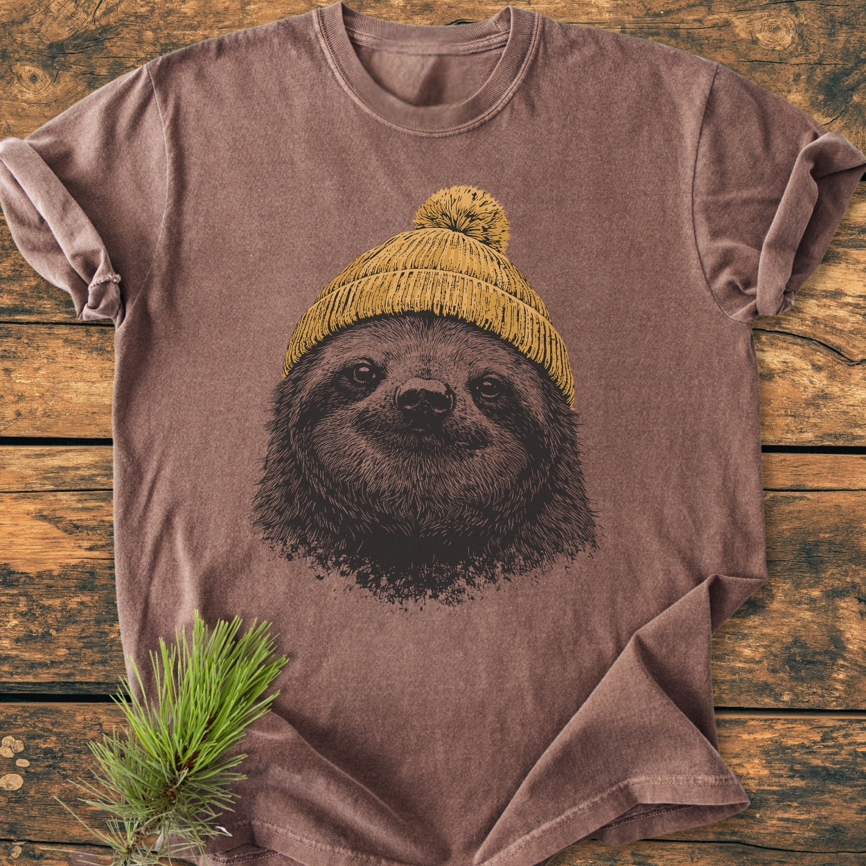 Sloth With Beanie - Vintage Wash Tee features a sloth face sketch wearing a knit beanie, displayed on a wooden surface with a pine sprig, highlighting its cozy, relaxed fit and durability.