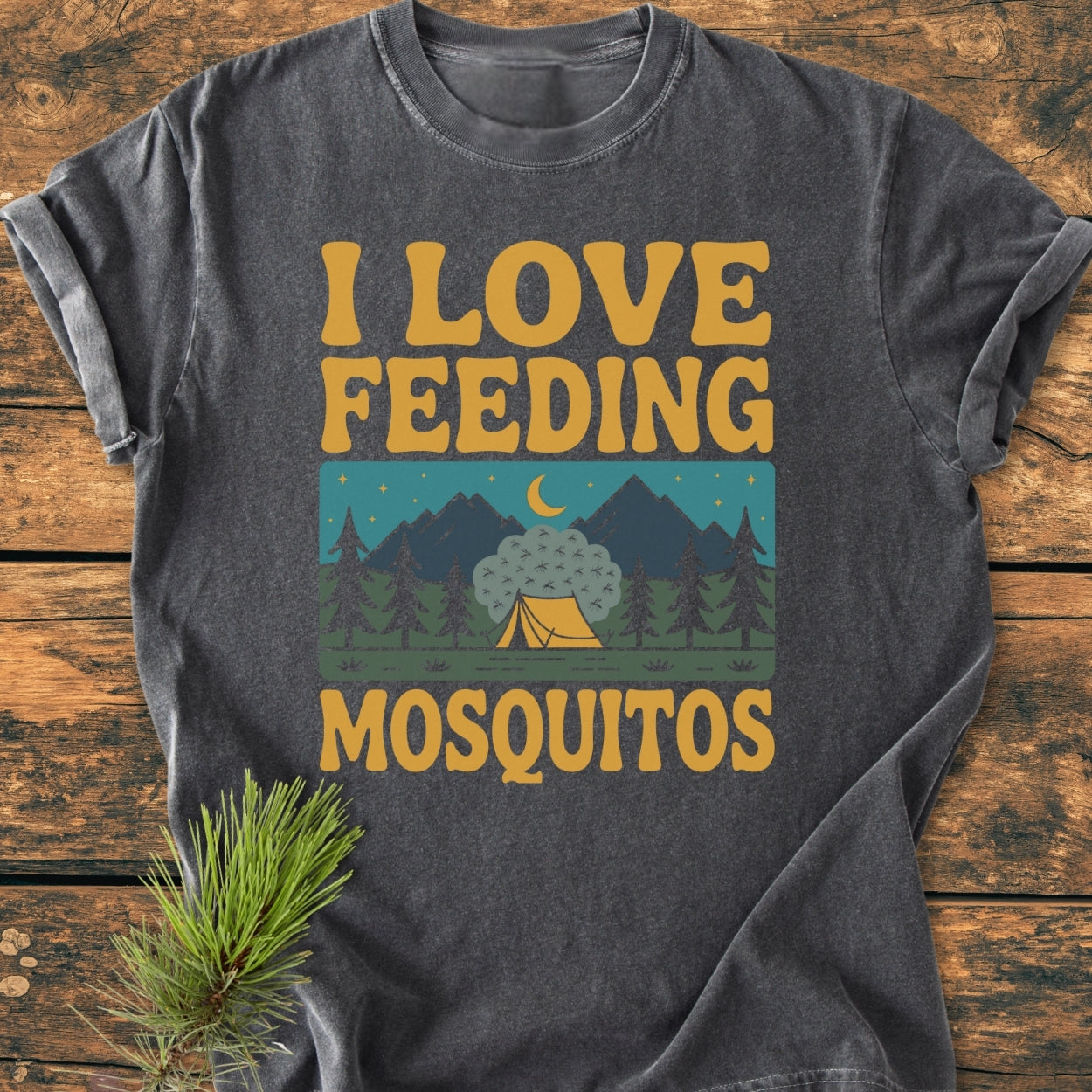 I Love Feeding Mosquitos - Vintage Wash Tee features a retro camping graphic with pine trees, mountains, and a moonlit tent, all on a dark-heather base, showcasing a playful mosquito-themed slogan.
