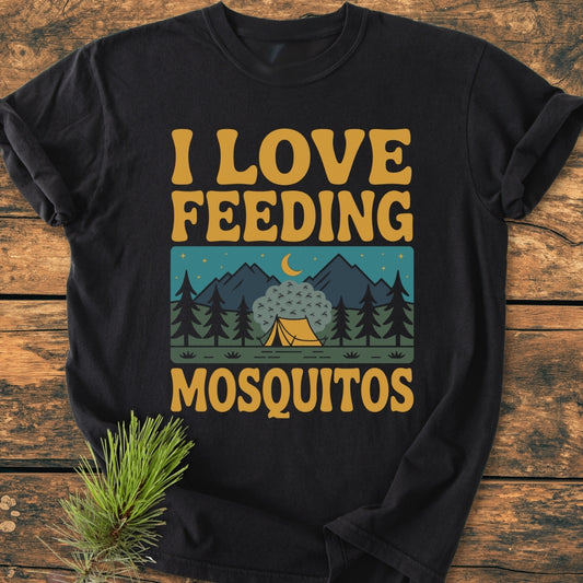 I Love Feeding Mosquitos - Vintage Wash Tee featuring mustard-yellow text and a retro camping scene illustration, displayed flat on wood. Includes evergreen sprig prop, showcasing the tee's relaxed fit and soft, durable fabric.