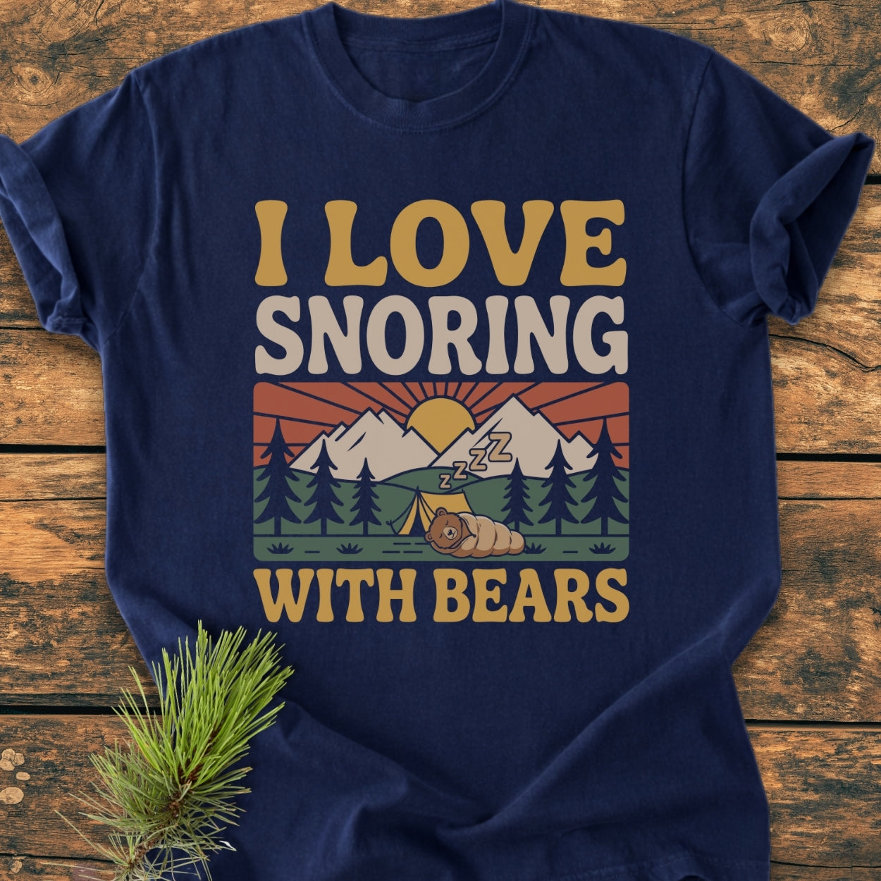 Navy-blue I Love Snoring With Bears - Vintage Wash Tee featuring a retro camping graphic with a cartoon bear, tent, and sunrise. Displayed on rustic wooden surface with a pine sprig accent.
